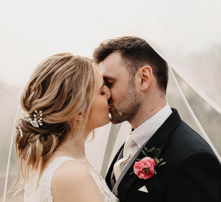 Groom in a dark morning suit and pink flower buttonhole shares a kiss with the bride 