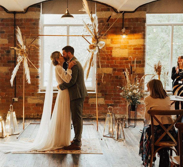 Bride & groom kiss in front of DIY brass arch complete with dried floral installations 