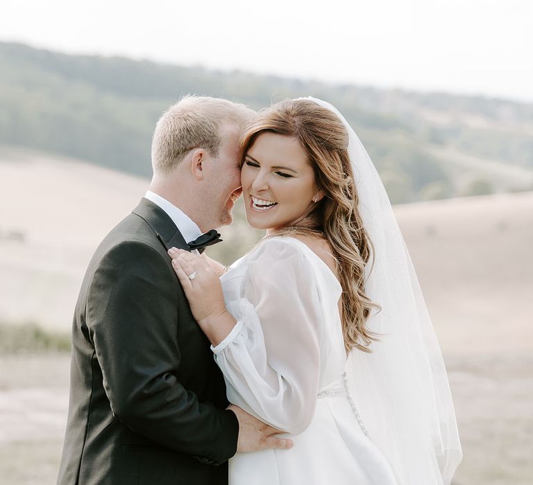 Bride in cathedral veil and sheer full sleeved wedding dress embrace outdoors for couples portraits