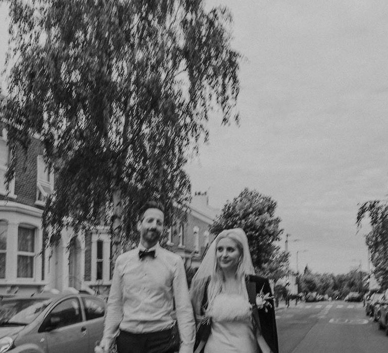 Bride in short satin dress with feather trimmed neckline and groom's blazer and groom in white shirt with bowtie holding hands and walking at Hackney Studios wedding