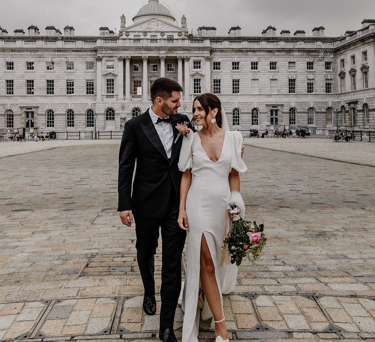 Bride in modern puff sleeve v neck wedding dress with front slit holding neutral floral bouquet and groom in dark tux walking around Somerset House