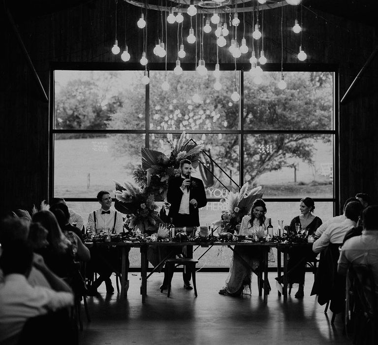 Groom stands up to read out his wedding speech standing in front of dried flower decor with festoon lighting 