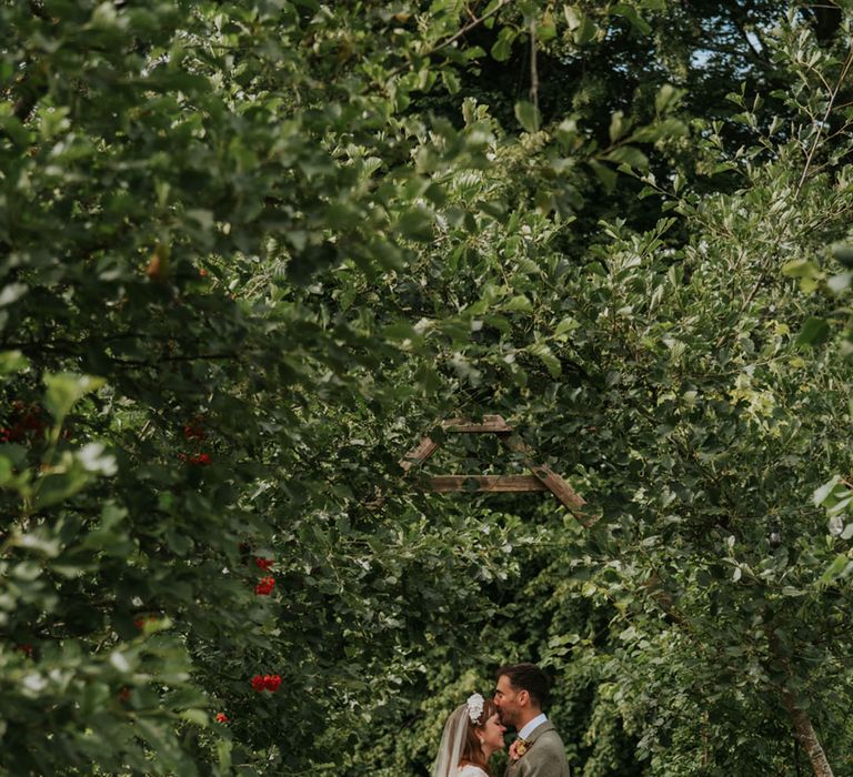 The groom in a tweed suit kisses the bride on the forehead as they pose for their couple portraits 