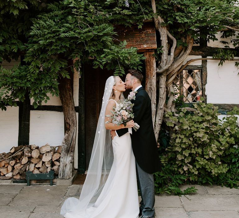 Bride in flower applique veil and simple wedding dress kisses the groom in three piece morning suit 