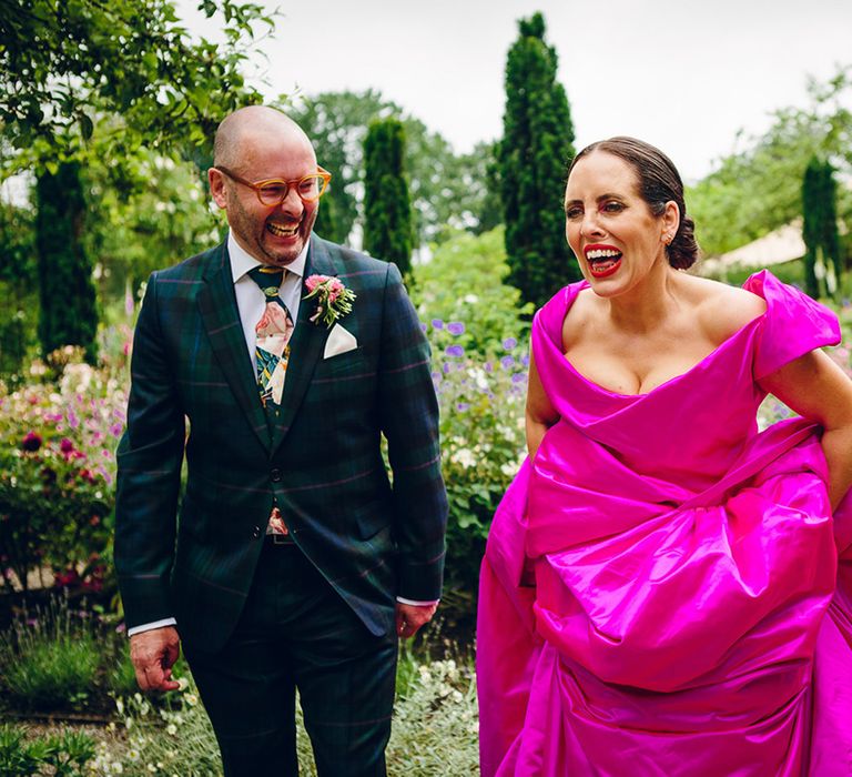 Groom in dark forest green tartan suit with patterned tie, white pocket square and pink and green boutonniere laughing with bride in off the shoulder front ruching pink Vivienne Westwood wedding dress