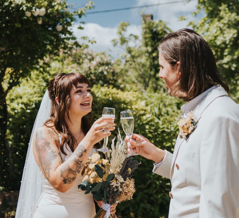 Bride in ASOS wedding dress with gold halo headband with sparkly stars having a glass of champagne with groom in neutral coloured double-breasted ASOS blazer