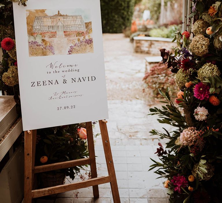 Wedding welcome sign with minimal type and watercolour painting stood on wooden easel by large floral wedding arch with white and pink carnations, garden roses, red strawflower, eucalyptus, baby’s-breath and various dried flowers 