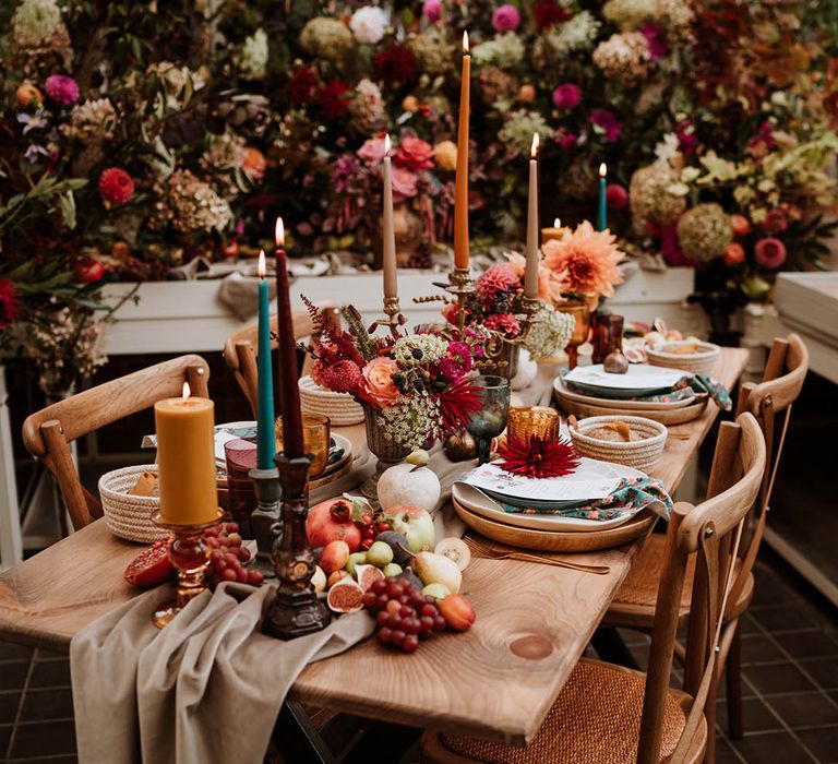 Neutral-toned tablerunner on rustic wooden table, orange, green and red coloured glasses, earthenware, seasonal fruit and vegetables, red, brown and green tapered candles in gold candlesticks and clear vases with autumnal coloured flowers with lots of roses, carnations, eucalyptus and other dried flowers in the background