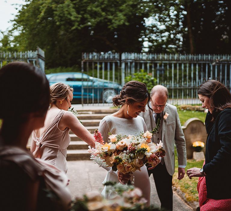 Wedding guests and bridesmaids in light pink bridesmaid dresses helping bride in long satin wedding dress holding tropical wedding bouquet with pink garden roses, foliage and baby's-breath 