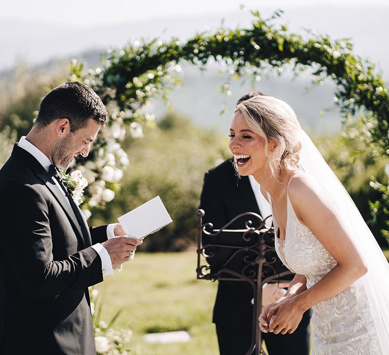 Blonde bride in lace wedding dress smiles as groom reads personal vows during outdoor wedding ceremony  