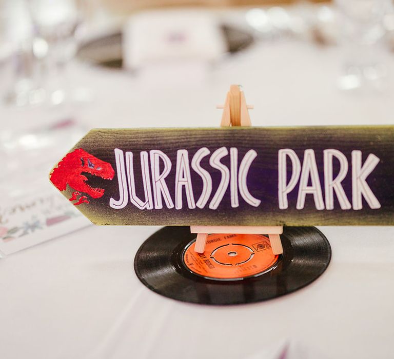 Quirky wedding table number sign in the style of Jurassic Park; cream arrow sign with "Jurassic Park" lettering on top of miniature vinyl record 