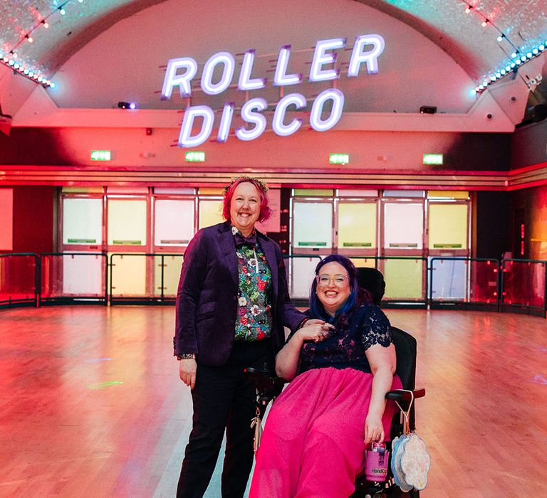 Bride in long pink wedding dress with purple bridal coverup on top with bride in purple velvet blazer and black suit trousers with purple bowtie, pink patterned pocket square and colourful patterned shirt in the roller disco of Margate Dreamland standing in front of large neon roller disco sign 