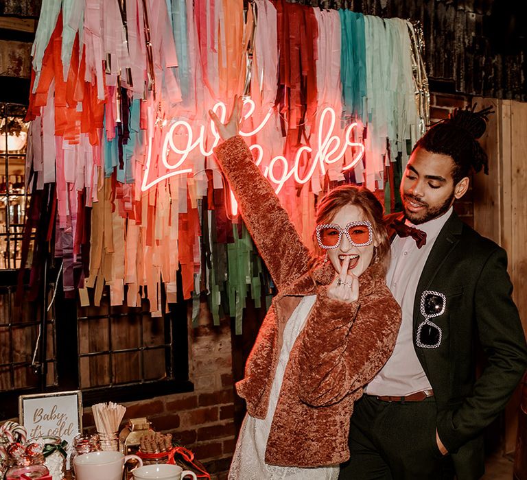 Bride in long sleeve lace wedding dress with caramel faux fur teddybear coat and large 60's-inspired pink wedding sunglasses posing with groom in classic black suit with burgundy bowtie posing in front of colourful wedding streamers and pink neon wedding sign 