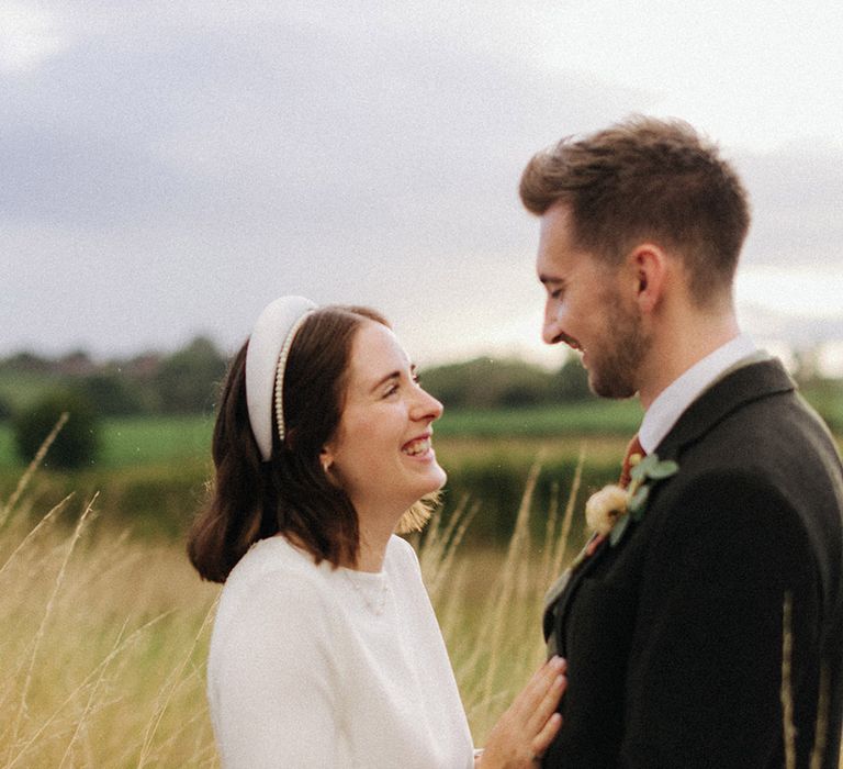 Laughing bride with short brown hair wearing a high neck three quarter length sleeves smiling at the groom in a field for their East Midlands wedding 