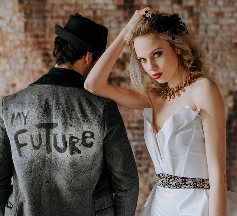 Groom in personalised dark grey jacket with "my future" graffiti style lettering and black fedora, and bride in structured bandeau wedding dress with partnered black belt, chunky brass choker necklace and black and white bridal hair accessories 