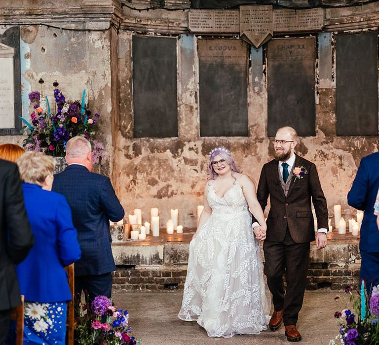 Bride in dainty leaf 3d appliqué sheer bridal gown and cathedral length veil holding hands with groom in mocha brown textured suit with deep green tie and pink and purple boutonniere walking down the aisle after bespoke wedding ceremony 