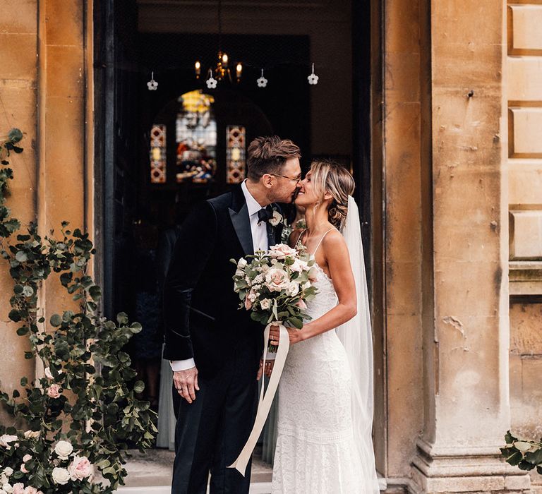 The bride wears a Grace Loves Lace spaghetti strap lace wedding dress leaning in to kiss the groom in a black tuxedo holding a pink rose bouquet 