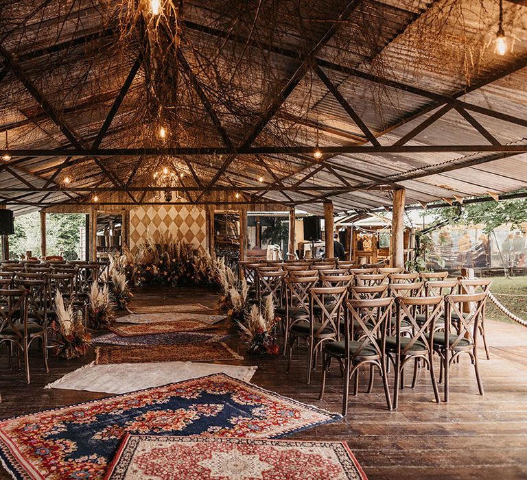 Barn wedding ceremony with Moroccan rugs and dried floral arrangements lining the aisle alongside wooden chairs 