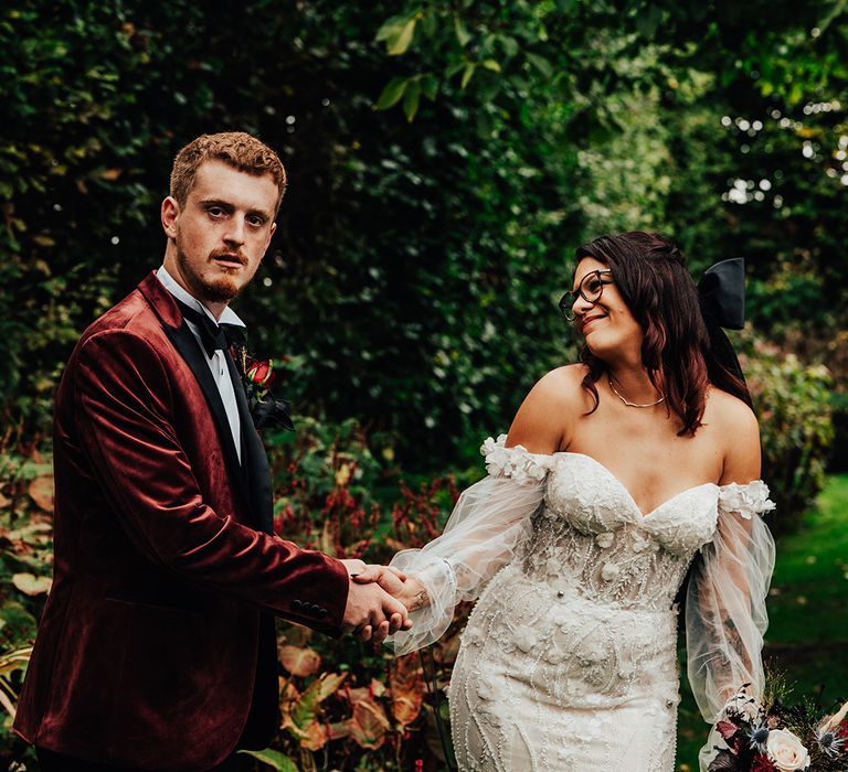 Groom in a burgundy suit with the bride in an off the shoulder floral beaded dress, glasses, and large black hair bow with skull crest 