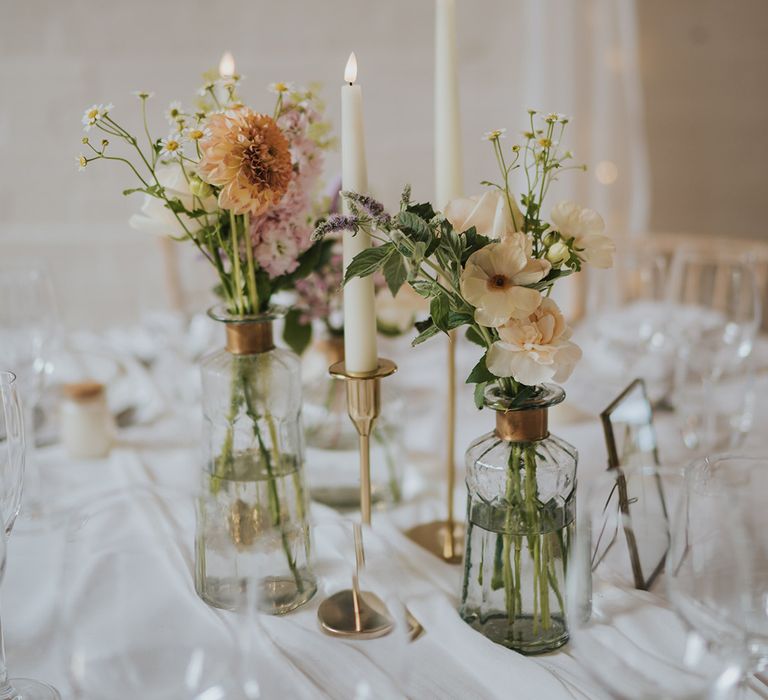 Glass bud vases with white candles in gold holders decorating the white tablescape with white tablecloth and table runner