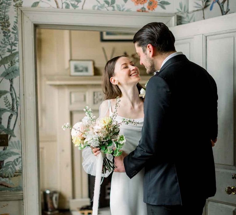 Bride in long sheer puff sleeve satin wedding dress with cowl neck holding classic bridal bouquet embracing groom in classic black tuxedo 