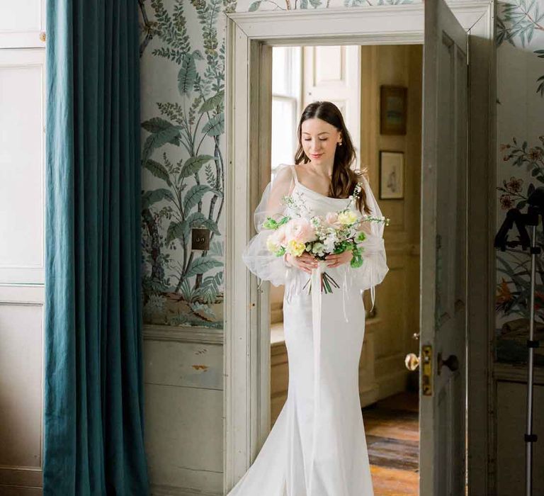 Bride standing in the doorway holding classic spring bouquet wearing cowl neck satin wedding dress with sheer puff sleeves 