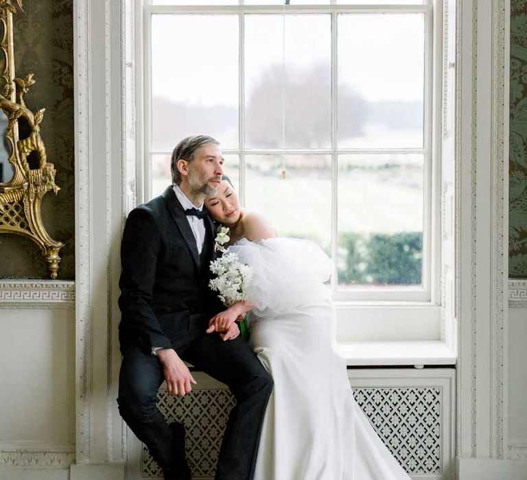 Groom in classic black tuxedo, black bowtie and boutonniere sitting with bride wearing strapless wedding dress with puff tulle sleeves