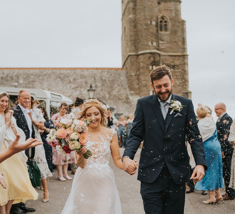 Bride in lace and tulle wedding dress holding pastel flowers walking with groom in navy suit for their confetti exit 