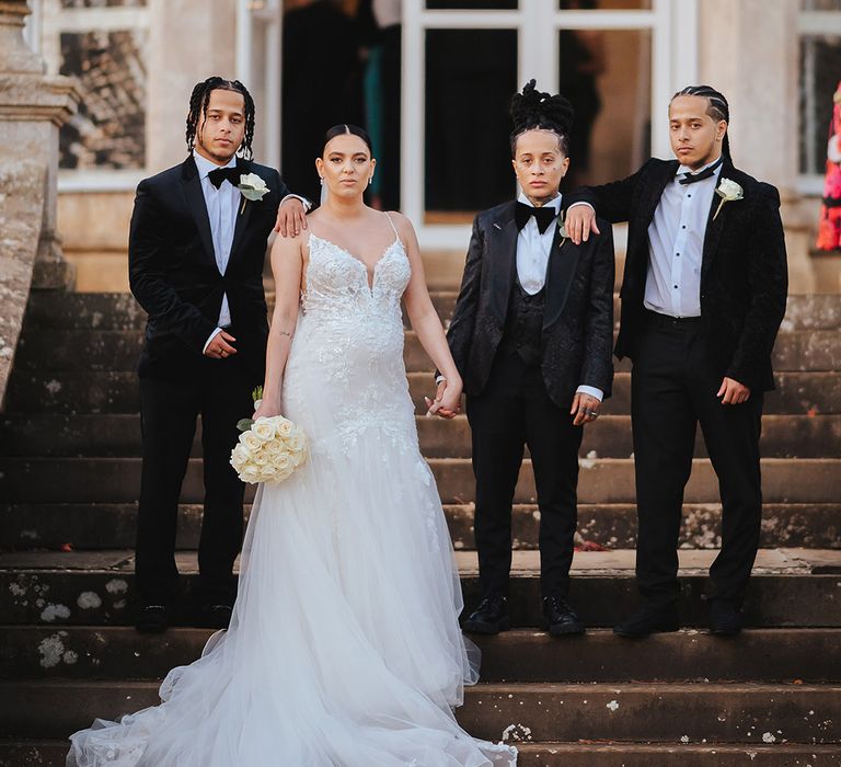 Bride in lace sleeveless v neck Enzoani wedding dress with tulle finish and church length veil holding hands with bride in black snakeskin print tuxedo with black bowtie and white rose boutonniere standing with bridal party in classic black tuxedos with white rose boutonnieres 