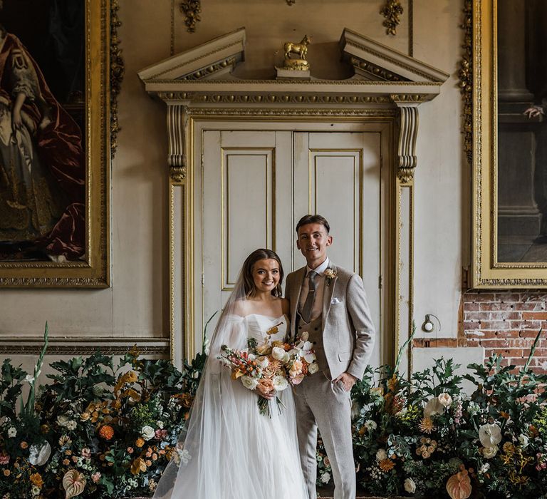 The bride in a whimsical wedding dress by Watters with the groom in a houndstooth beige suit surrounded by autumnal wedding flower arrangement 