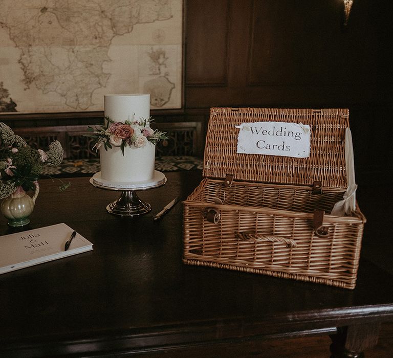 A wooden table with the two tier white iced wedding cake with pink and white rose decoration with a wicker basket for cards 