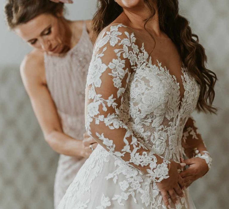 Mother of the bride in dusky pink sleeveless dress and dusky pink fascinator helping bride with long sleeve lace illusion sleeve wedding dress with overlay 