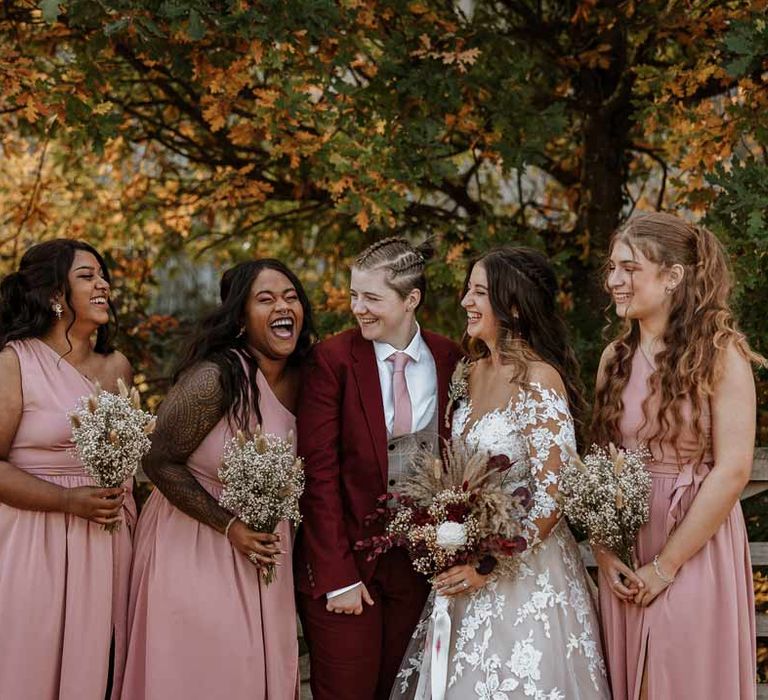 Bridesmaids in dusky pink one shoulder bridesmaid dresses standing with bride in maroon bridal suit with grey waistcoat, dusky pink tie and autumnal dried flower boutonniere and bride in long sleeve lace illusion sleeve wedding dress with overlay holding large autumnal boho dried flower bouquet 