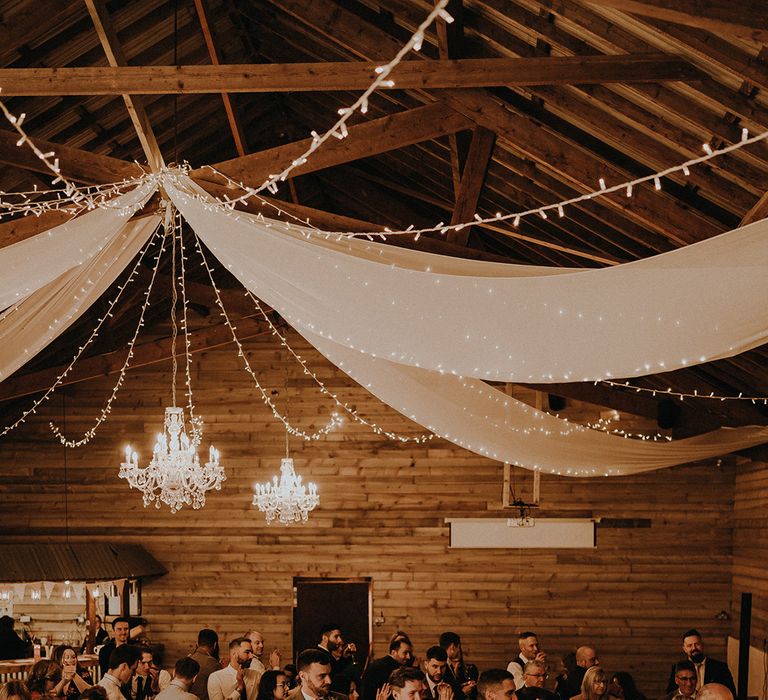 White drapery with chandeliers and fairy lights decorate the barn wedding venue with a rustic romance wedding theme 