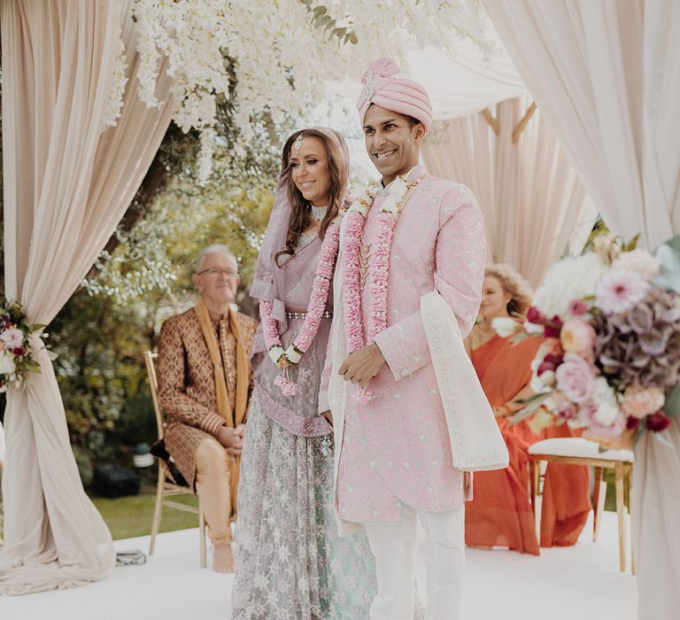 Groom wearing light pink sherwani, light pink turban and cream trousers and baby pink flower garland standing with bride in mint green and dusky pink ombre embellished lehenga and baby pink flower garland 