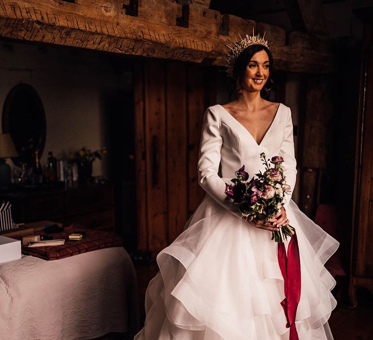 Bride in a layered ruffled wedding dress holding a dark pink winter bouquet tied with a red ribbon with gold crystal bride crown 