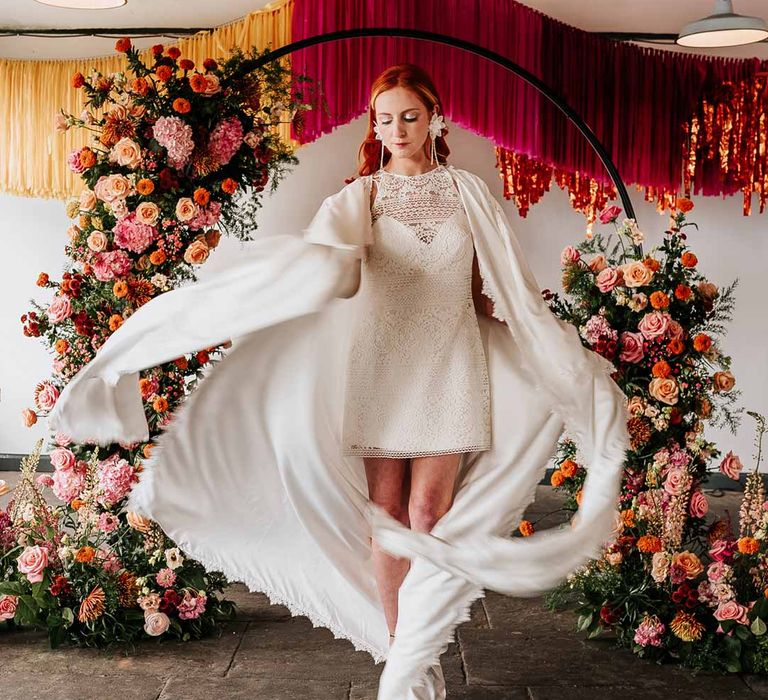 Bride in 70s-inspired boatneck short lace wedding dress with detachable tulle puff sleeves, platform gold wedding heels and scallop hem bridal cape at Northern Monk Refectory Leeds standing by circular floral arch with vibrant flowers and foliage attached and pink, red, yellow and metallic wedding streamers behind