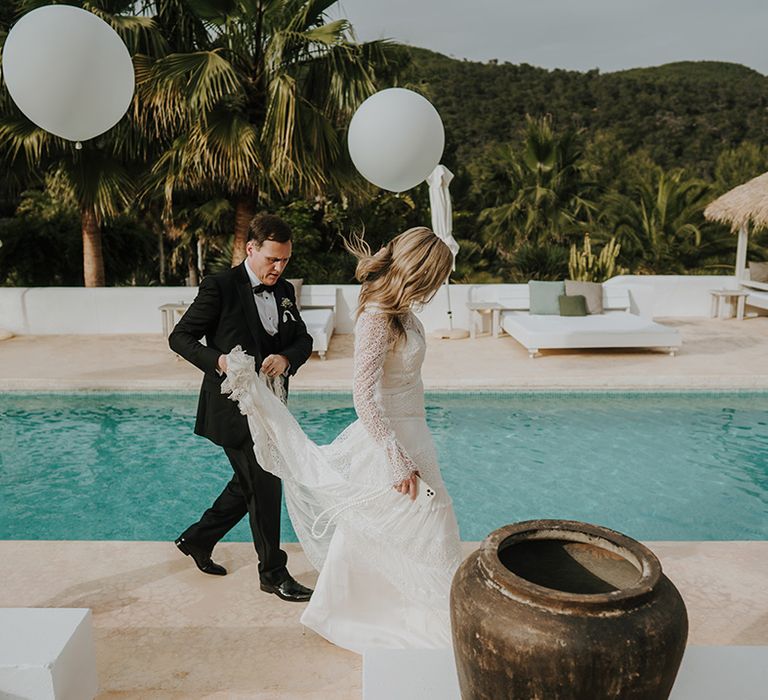 groom in a black tuxedo holding his brides wedding dress train as they walk past the pool at their pure house ibiza wedding