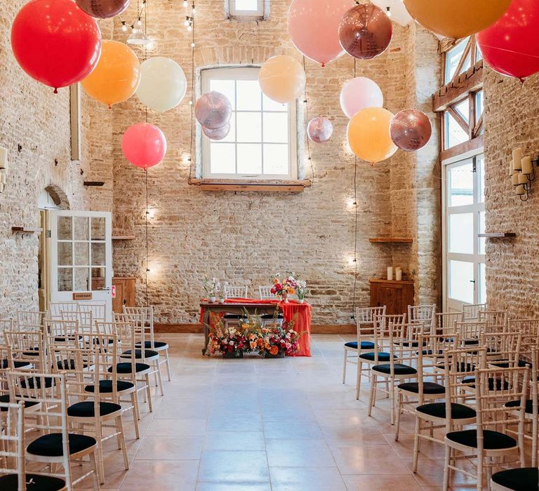 Merriscourt wedding venue reception room with white wooden chairs, metallic pink, white, orange and yellow balloons, wedding fairy lights decorations and wooden table with orange table runner and mixed seasonal flower arrangements 