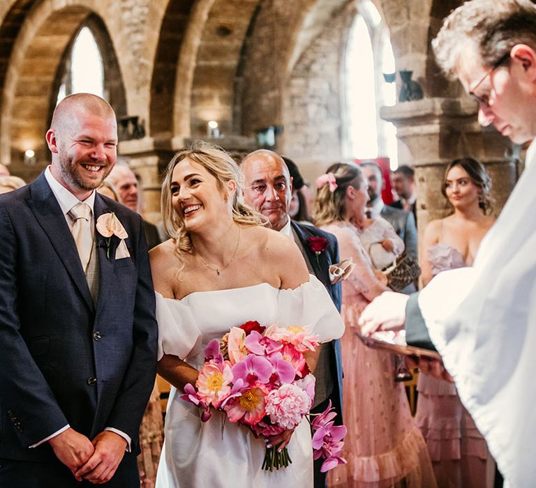 Groom in dark navy suit with pale pink anthurium buttonhole laughing with the bride at their church ceremony in the bride's hometown 