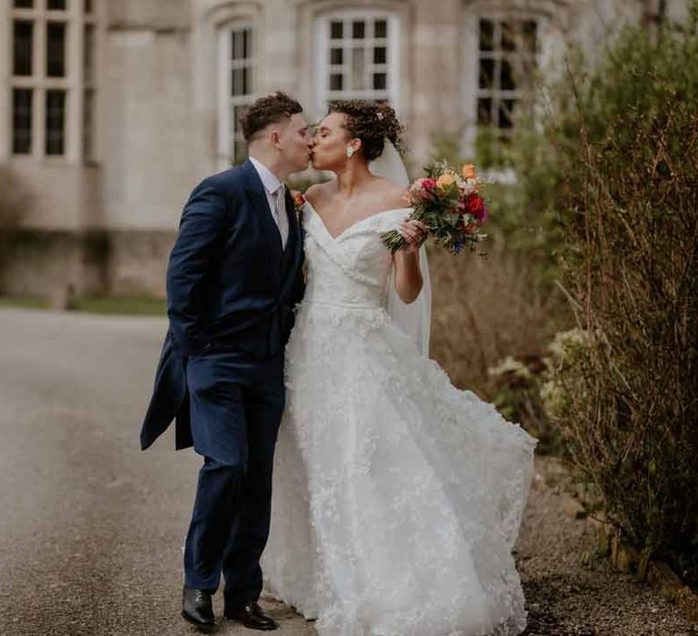 Groom in navy groom morning suit with grey ties and blush rose boutonniere kissing bride in Suzanne Neville off shoulder 3D floral applique wedding dress at Elmore Court Gloucestershire wedding venue