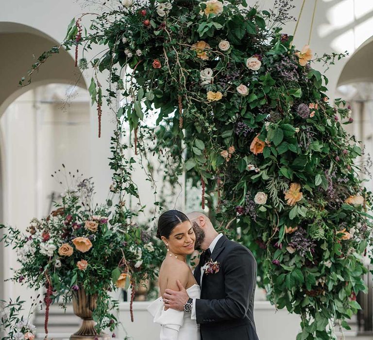 Groom in classic black tuxedo with bowtie and wildflower boutonniere embracing bride in long sleeve off the shoulder wedding dress with pearl button detailing and a side slit holding dried wildflower bouquet surrounded by suspended wildflower and foliage arrangements in large gold vases at Buxted Park