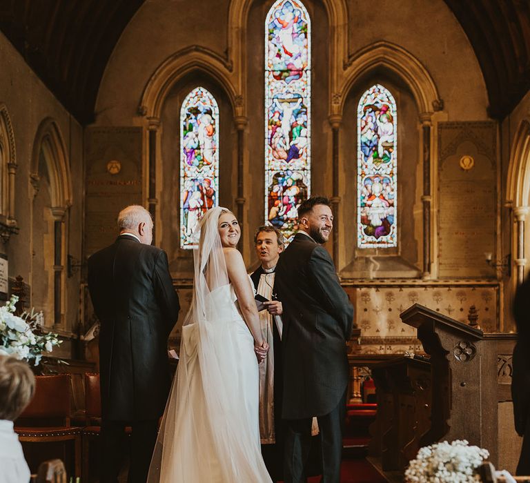 Bride in strapless wedding dress with bow design on the back with white veil looking back at the wedding guests with the groom at the altar 