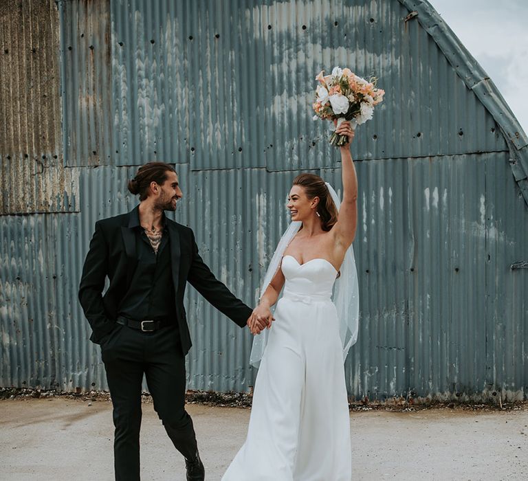 Bride in white bridal jumpsuit with pearl veil walking with groom in all black wedding suit 