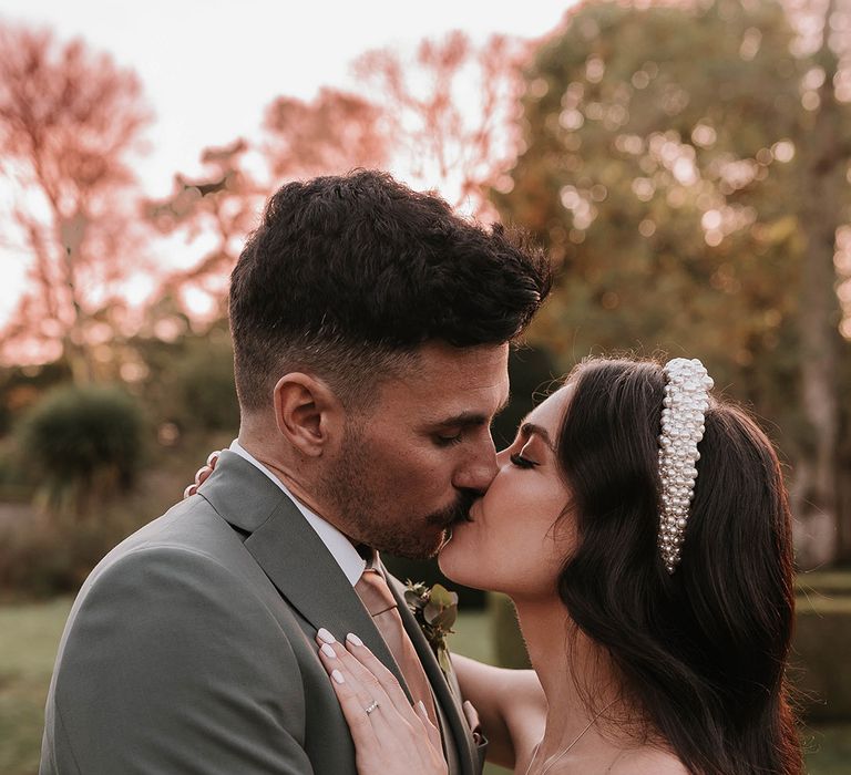 Bride wearing delicate pearl wedding headband posing with groom in green suit 