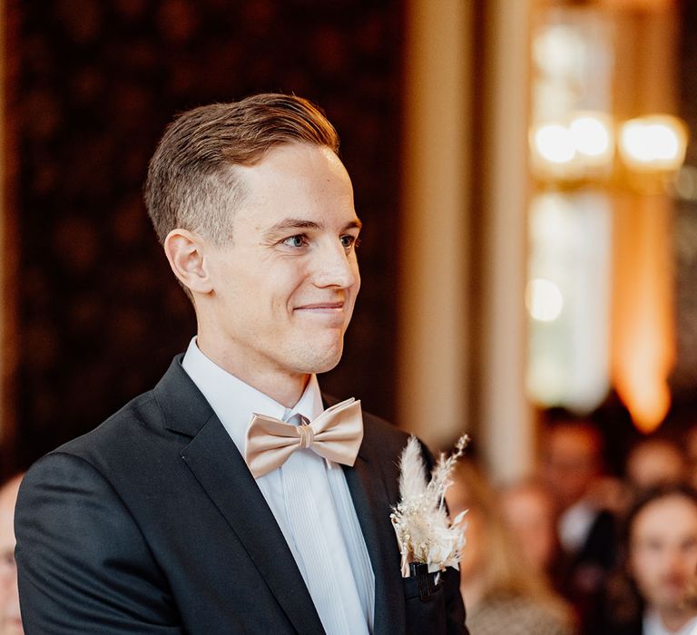 Groom in black Hugo Boss wedding suit with champagne bow tie and dried flower buttonhole smiling as he partakes in his wedding ceremony 