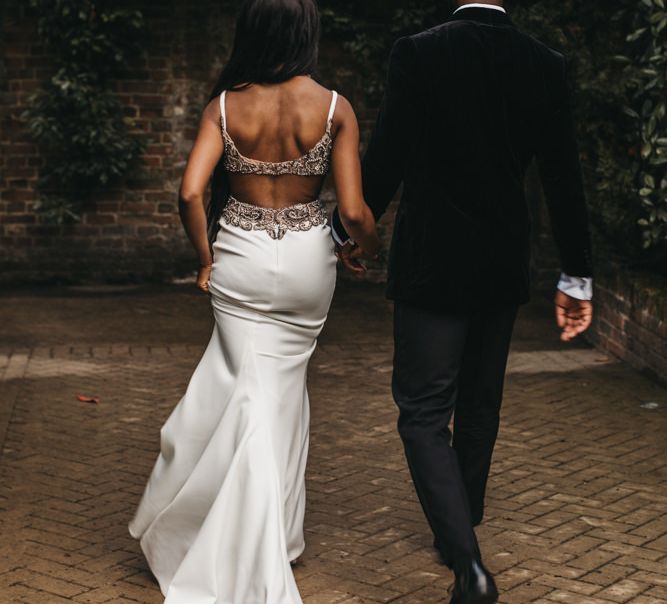 Black bride in a fitted wedding dress with low back detail and embellished straps holding her husbands hand in a velvet jacket 