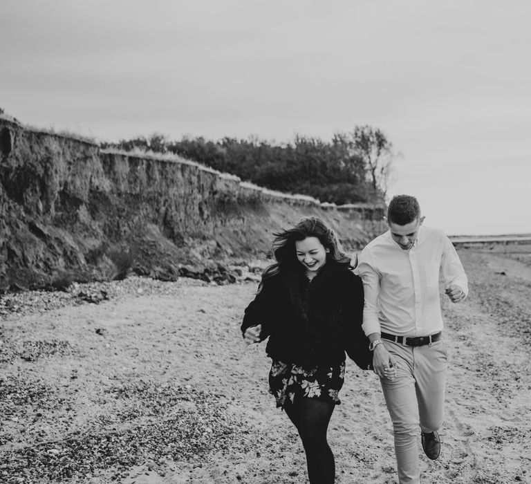 Bride-to-be in a floral dress and faux fur coat running on the beach with her groom-to-be