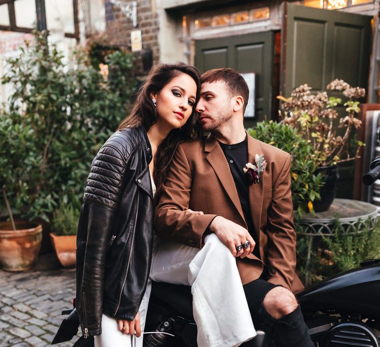 A bride and groom sit on a motorbike. She has brunette hair and a leather jacket draped over her shoulder. Her shoes are green with bows on the front and she wears white trousers. He wears a brown suit. The motorbike is black. Photography by Kirsty Mackenzie.