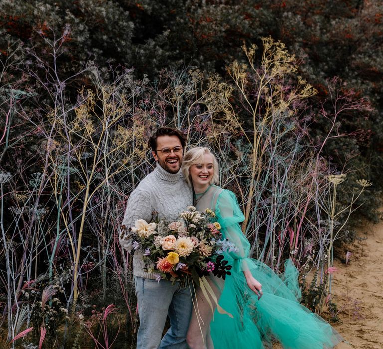 Barefoot bride and groom at their beach elopement standing in the sand by a colourful flower wall holding a bright wedding bouquet 